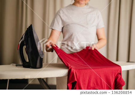 Woman ironing a long sleeve clothing at home. Daily chores. Laundry day. Neutral background. Home appliances 136208854