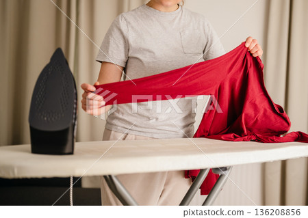 Woman ironing a long sleeve clothing at home. Daily chores. Laundry day. Neutral background. Home appliances Woman ironing a long sleeve clothing at home. Daily chores. Laundry day. Neutral background. Home appliances 136208856