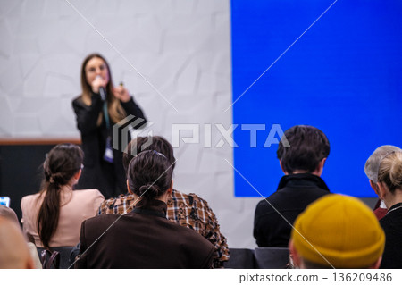 Speaker presenting to audience in conference setting with blue backdrop and attentive attendees Speaker presenting to audience in conference setting with blue backdrop and attentive attendees 136209486