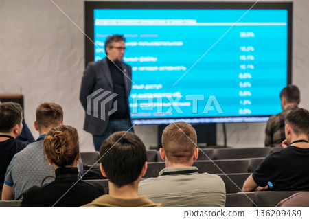 Professional conference presentation with speaker addressing audience in front of blue screen during lecture Professional conference presentation with speaker addressing audience in front of blue screen during lecture 136209489