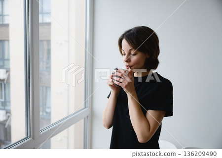 Woman drinking hot beverage by window at home Woman drinking hot beverage by window at home 136209490