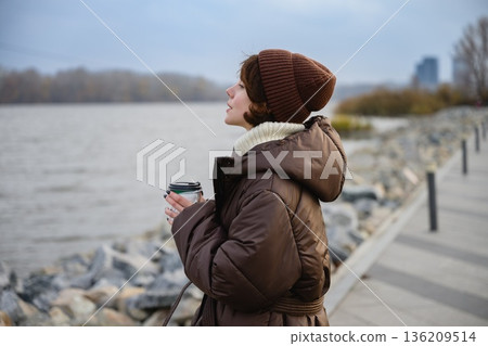 Woman in warm jacket holding coffee by river embankment Woman in warm jacket holding coffee by river embankment 136209514