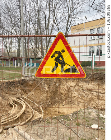 Caution sign indicating construction work ahead, featuring a worker with a shovel, surrounded by a fenced area with exposed earth and trees in the background, emphasizing safety awareness Caution sign indicating construction work ahead, featuring a worker with a shovel, surrounded by a fenced area with exposed earth and trees in the background, emphasizing safety awareness 136209868