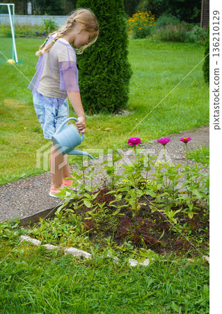 A smiling girl finds pleasure in gardening, gently showering her plants with water from a rustic watering can during a perfect summer day. 136210129