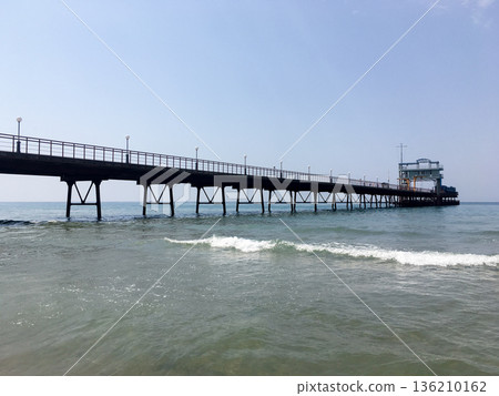 Long wooden pier stretches out over gentle waves of serene sea. Calm waters reflect bright blue of sky, peaceful atmosphere. Inviting beach location, perfect for relaxation and enjoying nature Long wooden pier stretches out over gentle waves of serene sea. Calm waters reflect bright blue of sky, peaceful atmosphere. Inviting beach location, perfect for relaxation and enjoying nature 136210162