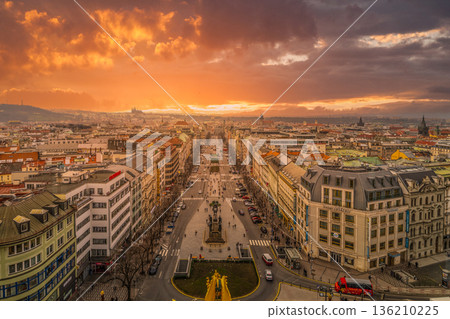 Wenceslas Square in Prague shows a wide view of the busy area as the sun sets. The sky is filled with colors and people walk along the street. Buildings line both sides of the square. 136210225