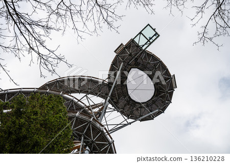 A wooden structure rises against the sky. People stand on the observation deck at the top. Cloudy weather surrounds the area and trees are viewed below. 136210228