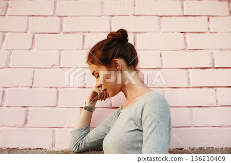 Young Woman Leaning On Pink Wall, Pensive Student Portrait Wearing Grey Sweater With Messy Bun And Wristwatch, Soft Natural Light, Muted Pastel Tones, Textured Brick Backdrop, Downward Gaze Conveying 136210409