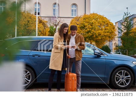 Man and woman with suitcase standing by car and checking smartphone Man and woman with suitcase standing by car and checking smartphone 136211073