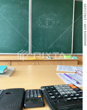 Classroom scene featuring a wooden desk with calculators, notebooks, and a chalkboard filled with mathematical equations and diagrams, creating an educational atmosphere for learning Classroom scene featuring a wooden desk with calculators, notebooks, and a chalkboard filled with mathematical equations and diagrams, creating an educational atmosphere for learning 136211285