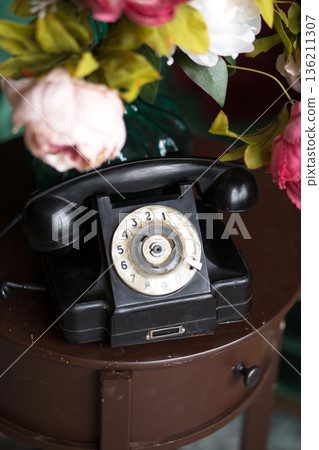 Vintage black rotary telephone placed on a round wooden table surrounded by colorful artificial flowers, creating a nostalgic and charming atmosphere for home decor 136211307
