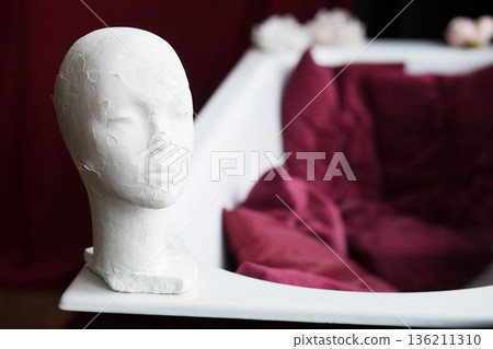 White sculpted head displayed beside a luxurious bathtub with deep burgundy fabric, creating an artistic and serene atmosphere in a contemporary interior design setting White sculpted head displayed beside a luxurious bathtub with deep burgundy fabric, creating an artistic and serene atmosphere in a contemporary interior design setting 136211310
