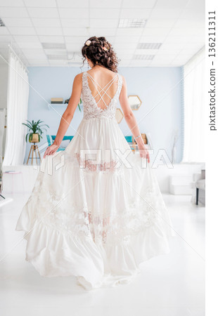 Elegant woman in a flowing white dress with intricate lace details stands gracefully in a bright, modern interior, showcasing beauty and style in a serene atmosphere Elegant woman in a flowing white dress with intricate lace details stands gracefully in a bright, modern interior, showcasing beauty and style in a serene atmosphere 136211311