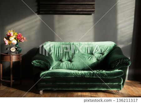 Retro interior featuring a plush green velvet sofa illuminated by soft sunlight, complemented by a vintage side table with flowers, creating a warm and inviting atmosphere 136211317