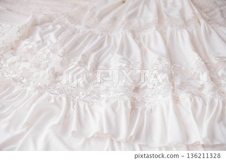 Elegant white bridal gown with intricate lace detailing and soft ruffles, beautifully arranged on a textured surface, showcasing delicate craftsmanship and romantic design elements Elegant white bridal gown with intricate lace detailing and soft ruffles, beautifully arranged on a textured surface, showcasing delicate craftsmanship and romantic design elements 136211328