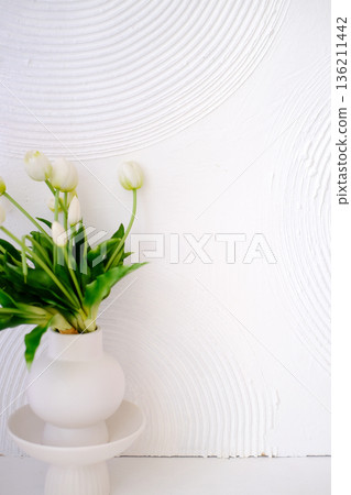 White vase with fresh tulips and green leaves placed on a minimalist pedestal against a textured white wall, creating a serene and elegant home decor atmosphere 136211442