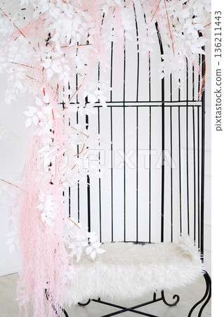 Elegant decorative birdcage adorned with white leaves and pink accents, featuring a plush fur seat, creating a serene and inviting atmosphere for interior design 136211443