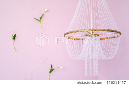 Elegant chandelier with delicate beadwork hangs from ceiling against soft pink wall, surrounded by minimalistic floral accents, creating a serene and stylish atmosphere. Copy space 136211457
