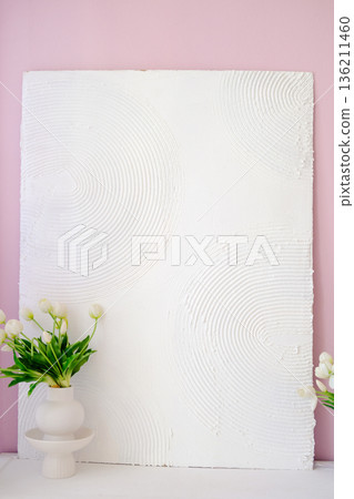 Textured white wall art with circular patterns, complemented by a minimalist vase of fresh flowers, creating a serene and stylish interior decor atmosphere. Copy space Textured white wall art with circular patterns, complemented by a minimalist vase of fresh flowers, creating a serene and stylish interior decor atmosphere. Copy space 136211460