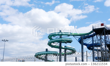 Empty water park featuring colorful water slides against a bright blue sky with fluffy clouds, creating a serene and tranquil atmosphere with ample copy space for design 136211534