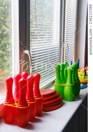 Colorful sports equipment for preschool children arranged on a windowsill, featuring red bowling pins and green cones, promoting active play and physical development in kindergarten settings 136211560