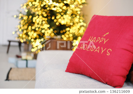 Cozy living room scene featuring a red decorative pillow with "Happy Holidays" text, soft sofa, and a beautifully lit Christmas tree in the background creating a festive atmosphere Cozy living room scene featuring a red decorative pillow with "Happy Holidays" text, soft sofa, and a beautifully lit Christmas tree in the background creating a festive atmosphere 136211609