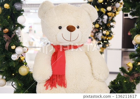 Large plush teddy bear with a red scarf is surrounded by festive decorations, creating a cheerful atmosphere for holiday celebrations and joyful moments Large plush teddy bear with a red scarf is surrounded by festive decorations, creating a cheerful atmosphere for holiday celebrations and joyful moments 136211617