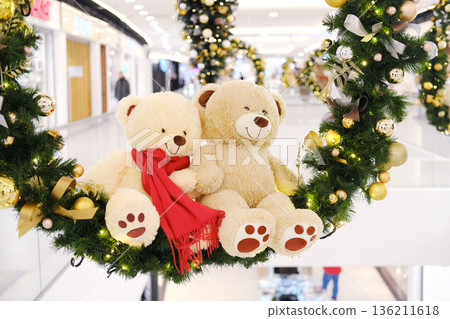 Two plush teddy bears adorned with festive scarves are nestled in a vibrant Christmas wreath, surrounded by sparkling ornaments in a bustling shopping center atmosphere Two plush teddy bears adorned with festive scarves are nestled in a vibrant Christmas wreath, surrounded by sparkling ornaments in a bustling shopping center atmosphere 136211618