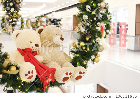 Two plush teddy bears with red scarf are nestled in a festive wreath adorned with ornaments, creating a joyful holiday atmosphere in a public shopping center Two plush teddy bears with red scarf are nestled in a festive wreath adorned with ornaments, creating a joyful holiday atmosphere in a public shopping center 136211619