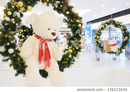 Plush teddy bear wearing red scarf hangs from festive wreath adorned with ornaments, creating a cheerful holiday atmosphere in a bustling shopping center Plush teddy bear wearing red scarf hangs from festive wreath adorned with ornaments, creating a cheerful holiday atmosphere in a bustling shopping center 136211639