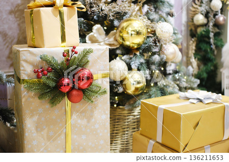 Festively wrapped gift boxes adorned with shiny ornaments and greenery, placed near a beautifully decorated Christmas tree, creating a warm holiday atmosphere Festively wrapped gift boxes adorned with shiny ornaments and greenery, placed near a beautifully decorated Christmas tree, creating a warm holiday atmosphere 136211653