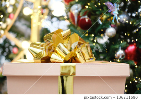 Gift box with golden ribbon and bow, placed in front of a beautifully decorated Christmas tree, adorned with colorful ornaments and twinkling lights, creating a festive atmosphere 136211680
