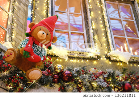 Plush teddy bear wearing a festive red hat and scarf is perched on a beautifully decorated garland with sparkling lights in a vibrant shopping center during the holiday season Plush teddy bear wearing a festive red hat and scarf is perched on a beautifully decorated garland with sparkling lights in a vibrant shopping center during the holiday season 136211811