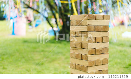 Wooden block tower stacked vertically on green grass with colorful ribbons hanging from tree branches in background during outdoor event with blurred objects nearby 136211839