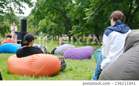 Group of individuals seated on colorful beanbags in a lush outdoor setting, attentively listening to a speaker during an engaging lecture with ample copy space 136211841