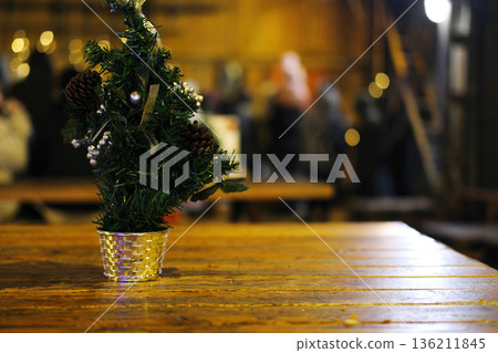 Small Christmas tree with pinecones and ornaments in a silver pot sits on a rustic wooden table, creating a festive atmosphere with blurred background lights Small Christmas tree with pinecones and ornaments in a silver pot sits on a rustic wooden table, creating a festive atmosphere with blurred background lights 136211845