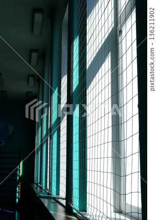 Bright sunlight streams through large windows with mesh netting, casting intricate shadows on the floor, creating a serene atmosphere in a modern indoor space Bright sunlight streams through large windows with mesh netting, casting intricate shadows on the floor, creating a serene atmosphere in a modern indoor space 136211902