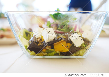 Fresh salad in a clear glass bowl featuring vibrant greens, colorful vegetables, and creamy feta cheese cubes, showcasing healthy eating and culinary presentation Fresh salad in a clear glass bowl featuring vibrant greens, colorful vegetables, and creamy feta cheese cubes, showcasing healthy eating and culinary presentation 136211919