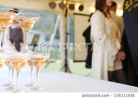 Champagne glasses stacked on white table with sparkling drink inside, blurred people in background, event setting with lighting and equipment visible in tent during daytime 136211936