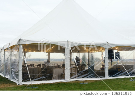 Outdoor celebration tent with transparent sides, elegantly decorated interior, providing shelter from weather elements, creating a festive atmosphere for gatherings and events Outdoor celebration tent with transparent sides, elegantly decorated interior, providing shelter from weather elements, creating a festive atmosphere for gatherings and events 136211955