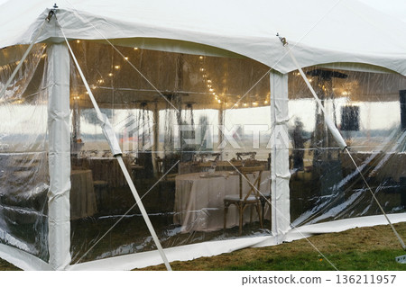 Clear tent with white interior and string lights, tables set with chairs inside, visible grass outside, during cloudy weather with no people present Clear tent with white interior and string lights, tables set with chairs inside, visible grass outside, during cloudy weather with no people present 136211957