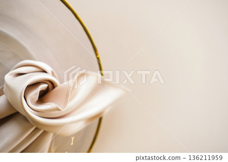 Wedding banquet table setting with white satin napkin tied in a knot on a gold-rimmed plate against a light beige background with copy space Wedding banquet table setting with white satin napkin tied in a knot on a gold-rimmed plate against a light beige background with copy space 136211959