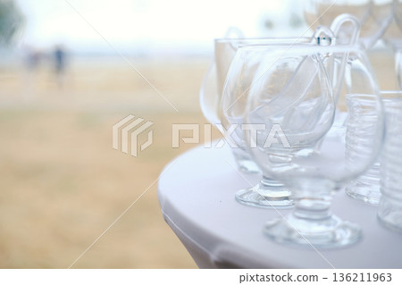 Clear glass cups arranged on white tablecloth with blurred background of outdoor setting and people walking in distance during daytime 136211963