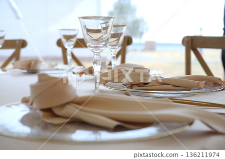 Table setting with beige napkins and glassware on round plates with gold trim, wooden chairs in background, soft lighting, and blurred outdoor view beyond the window Table setting with beige napkins and glassware on round plates with gold trim, wooden chairs in background, soft lighting, and blurred outdoor view beyond the window 136211974