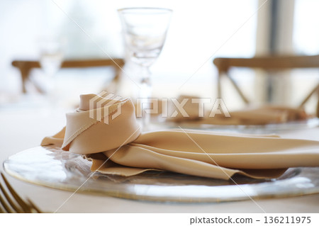 Table setting with beige napkin elegantly arranged on silver plate beside glassware and wooden chairs in bright indoor dining area with soft natural light Table setting with beige napkin elegantly arranged on silver plate beside glassware and wooden chairs in bright indoor dining area with soft natural light 136211975