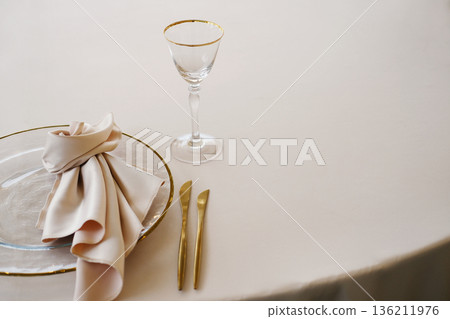 Wedding banquet table setting with white tablecloth, gold-rimmed plate, folded napkin, glass, and cutlery arranged neatly with copy space for design Wedding banquet table setting with white tablecloth, gold-rimmed plate, folded napkin, glass, and cutlery arranged neatly with copy space for design 136211976