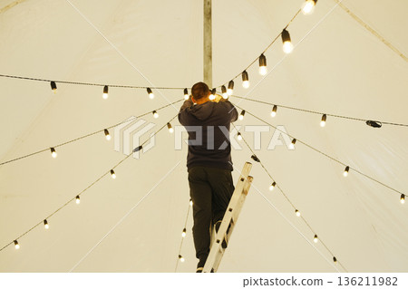 Male technician adjusting decorative string lights on a ladder inside a large tent, creating a warm ambiance for an outdoor event with illuminated atmosphere Male technician adjusting decorative string lights on a ladder inside a large tent, creating a warm ambiance for an outdoor event with illuminated atmosphere 136211982
