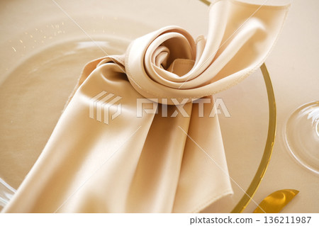 Elegant beige satin napkin artfully arranged on a glass plate, showcasing fine dining aesthetics, perfect for event planning and table setting inspiration 136211987