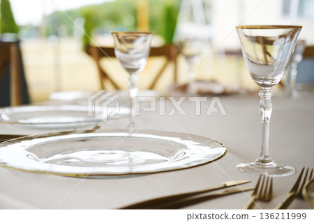 Elegant dining table setting with clear glass plates, crystal wine glasses, and gold-accented cutlery on beige tablecloth with blurred outdoor background and wooden chairs 136211999