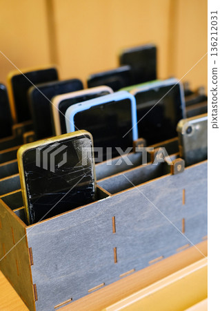 Collection of cell phones stored in a wooden box, showcasing various devices with cracked screens, representing the ban on mobile phones in educational environments and promoting focus in classrooms 136212031